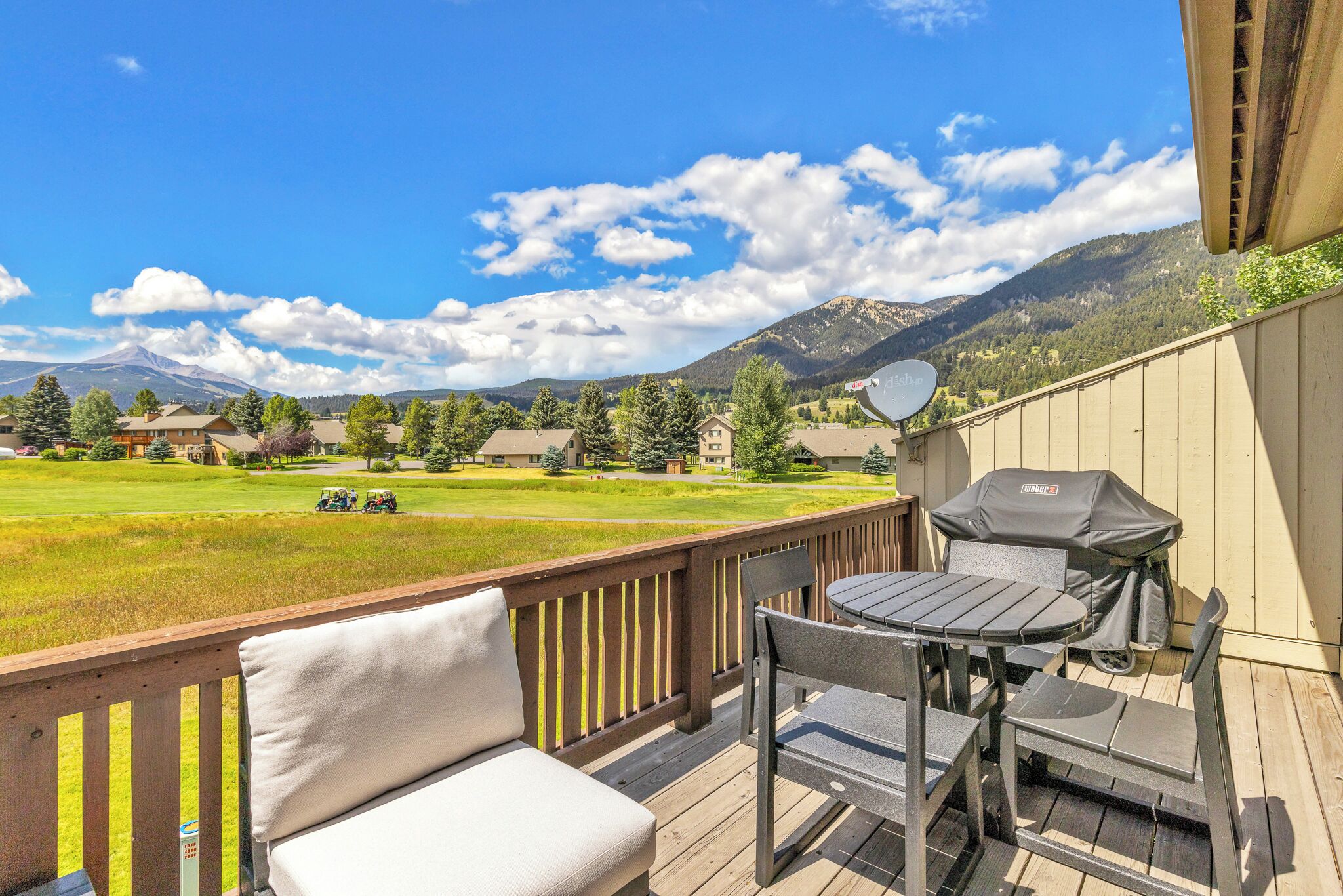 A deck with a grill, a table, and a chair, overlooking a green field and mountains.