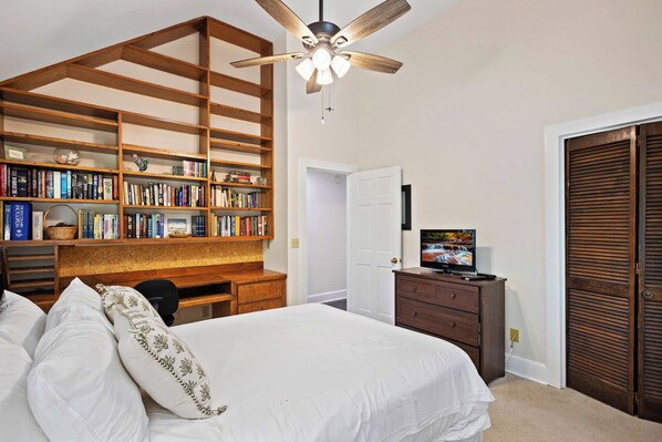 Ein Schlafzimmer mit einem Bett, einem Schreibtisch, einem Bücherregal, einem Deckenventilator, einem Fernseher und einem Kleiderschrank.