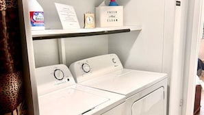 Une buanderie équipée de deux machines à laver à chargement frontal et d’un sèche-linge, d’une étagère contenant des produits de nettoyage et d’une porte donnant sur une autre pièce.