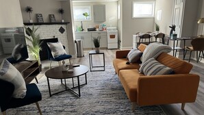 Un salon moderne avec un canapé, des fauteuils, une table basse et une cheminée. On aperçoit, au fond, un coin cuisine avec une table à manger et des chaises.