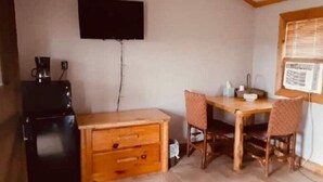 Una habitación compacta con un escritorio de madera, dos sillas, un refrigerador negro pequeño y un televisor de pantalla plana montado.