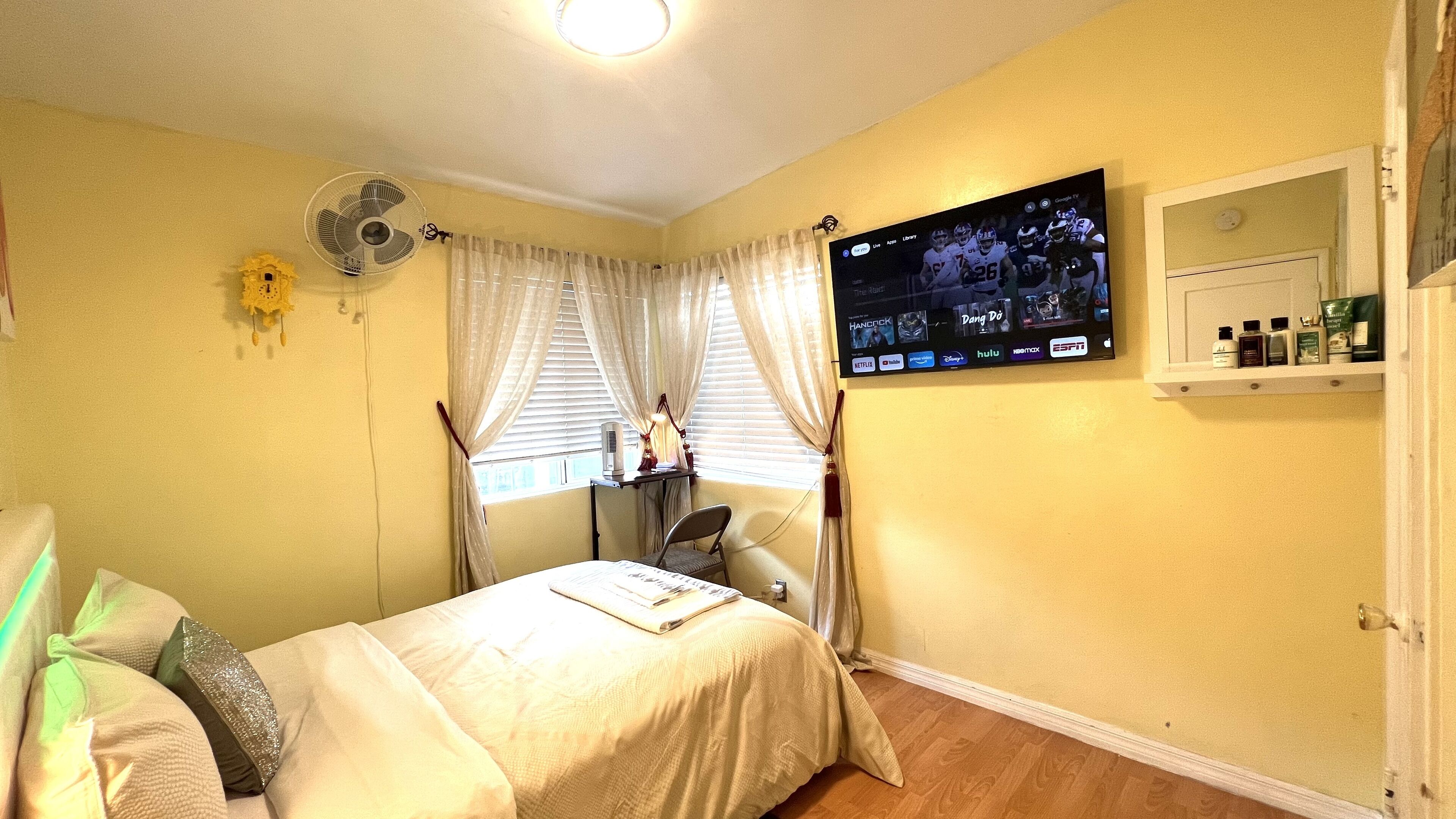 A bedroom with a bed, a desk, a TV mounted on the wall, and a window with curtains.