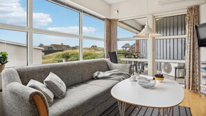 A cozy living room with a grey sofa, a round table with a bowl of fruit, and large windows offering a view of houses and greenery. - Amazing apartment in Fanø with WiFi (Fanø)