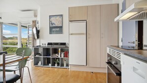 A modern kitchen with a built-in refrigerator, oven, and microwave. There is a dining area with a table and chairs, and a bookshelf with various items. - Amazing apartment in Fanø with WiFi (Fanø)