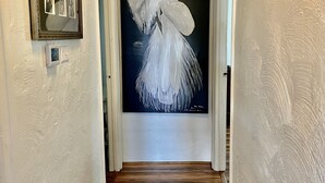 A hallway with a wooden floor, a framed photograph on the left, and a large painting of a bird on the right.