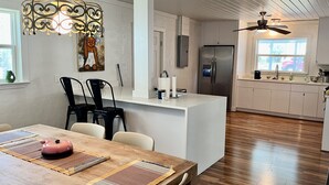 A modern kitchen with white cabinetry, a stainless steel refrigerator, a dining area with a wooden table and chairs, and a pendant light fixture.