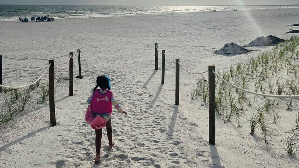 A sandy beach with a child walking towards the ocean, a rope barrier, and a clear sky.