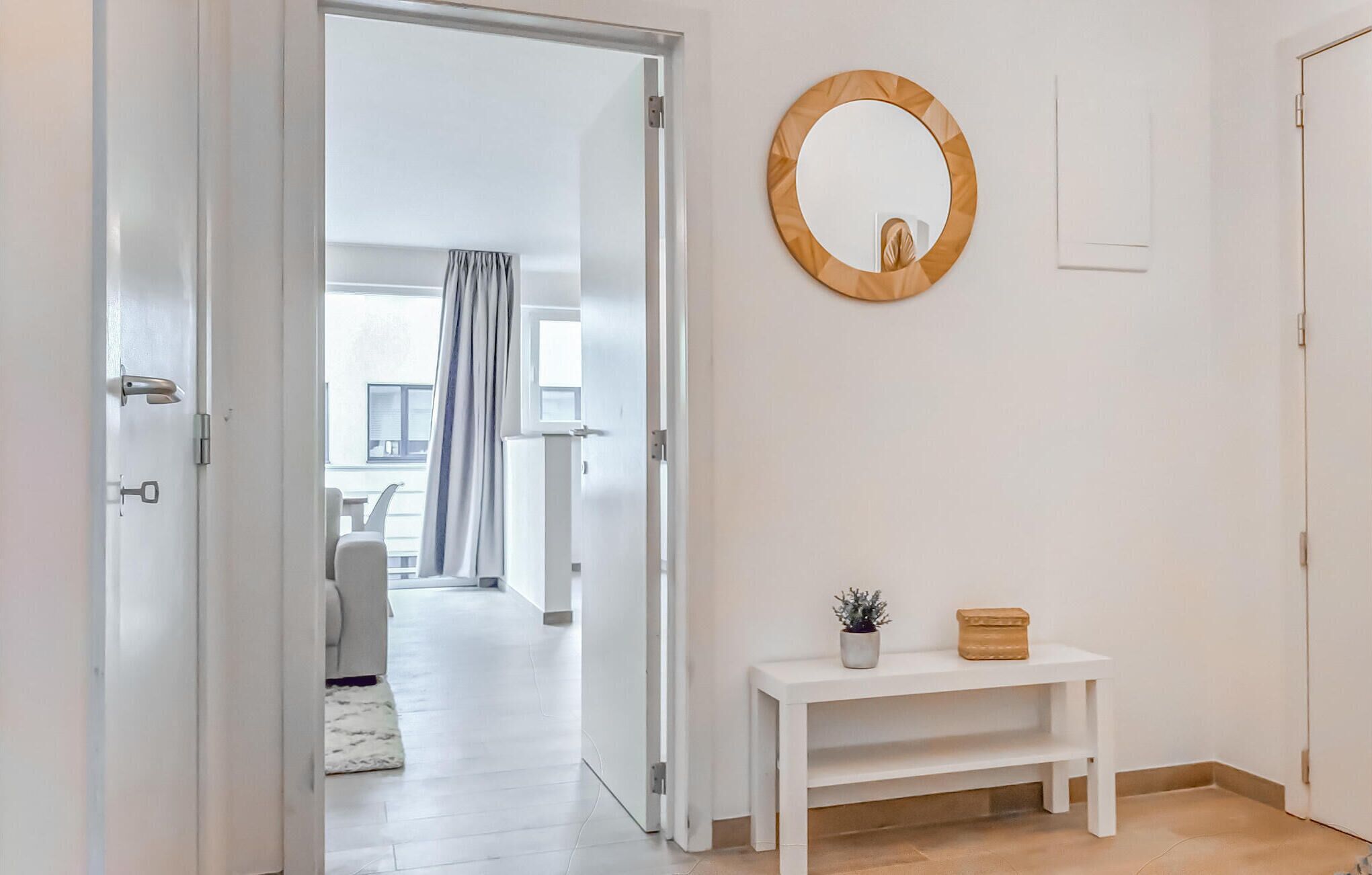 Um corredor com uma porta branca, um espelho redondo e um pequeno console branco com uma planta e uma caixa.