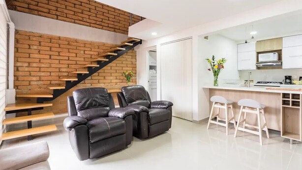 Un espacio habitable moderno con una escalera de ladrillo, sillones de cuero, una zona de cocina y una barra para comer.