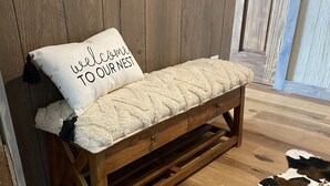 Un banco de madera con un cojín y una almohada que dice 'welcome to our nest' contra una pared de madera.
