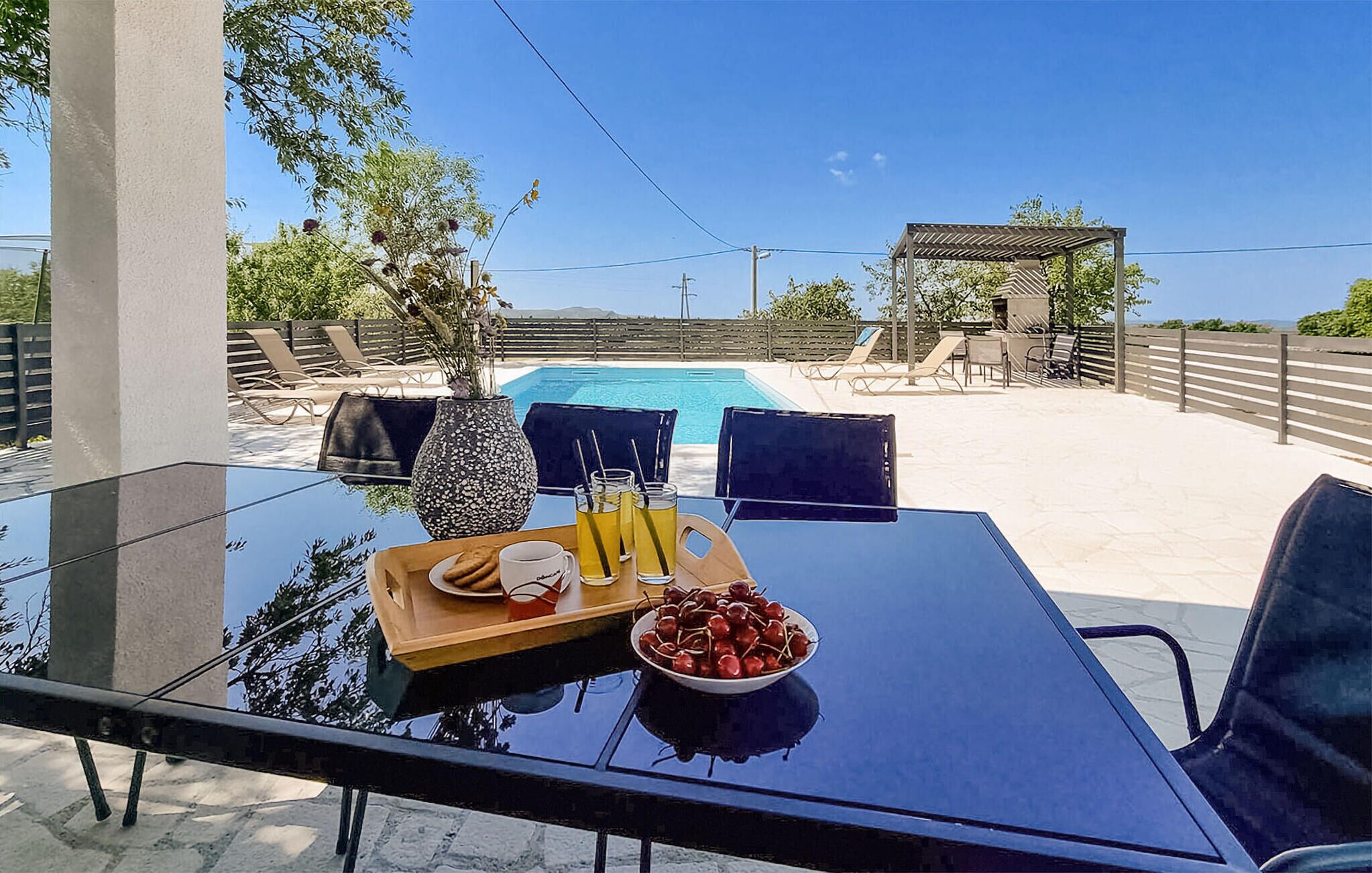 A glass table with a tray of food and drinks, a bowl of cherries, and a view of a swimming pool and outdoor seating area.