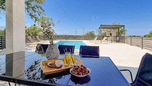 Ein Glastisch mit einer Speise- und Getränkeauswahl, einer Kirschschale und Blick auf einen Swimmingpool sowie einen Außenbereich mit Sitzgelegenheiten.