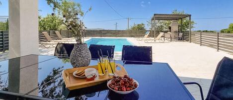 A glass table with a tray of food and drinks, a bowl of cherries, and a view of a swimming pool and outdoor seating area.