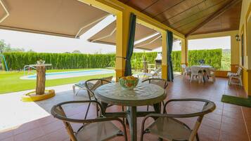Een overdekt terras met een ronde tafel en stoelen, een klein zwembad en uitzicht op een verzorgde tuin.