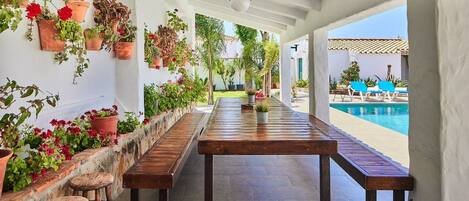 A covered patio with a wooden table and benches, surrounded by potted plants and a view of a swimming pool.