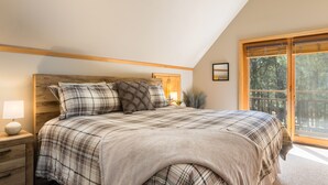 Camera da letto con testiera in legno, copripiumino a quadri, comodino con lampada e un'ampia finestra vista alberi.