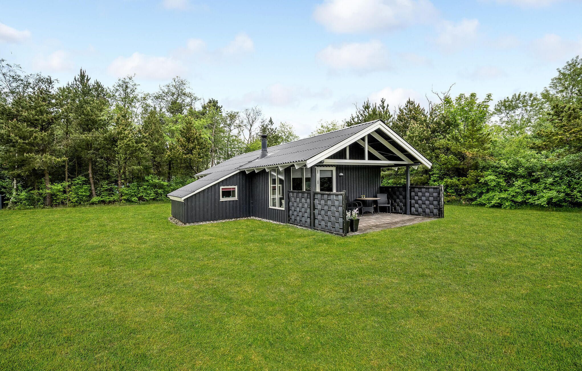 A small, dark-colored cabin with a sloped roof, a porch, and a fenced outdoor area.