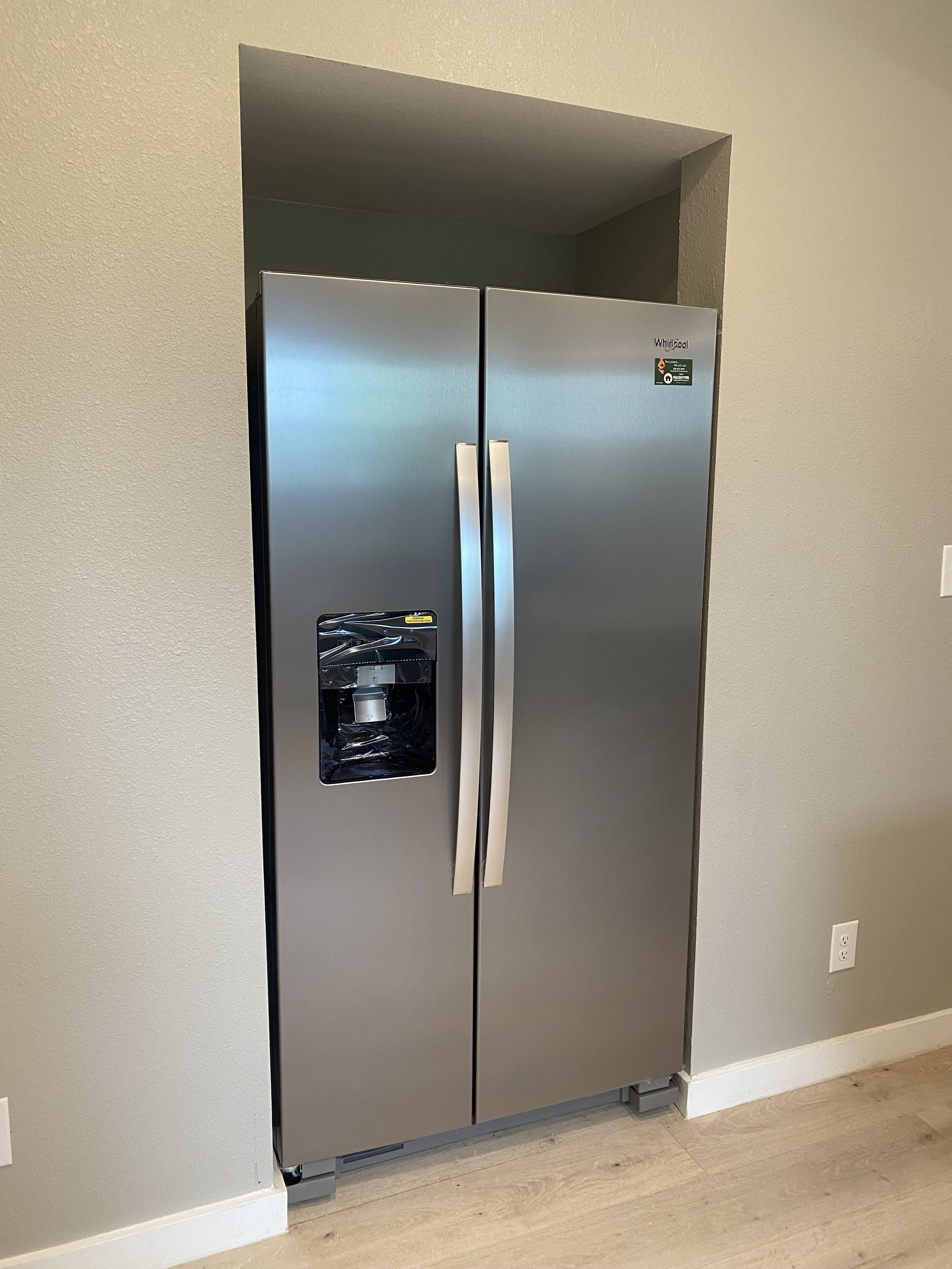 A stainless steel Whirlpool refrigerator with a water dispenser, located in a room with beige walls and wooden flooring.