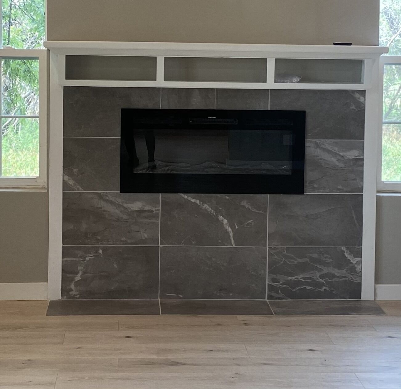 En moderne pejs med et fladskærms-tv over, i et rum med flisegulv og et vindue til venstre.