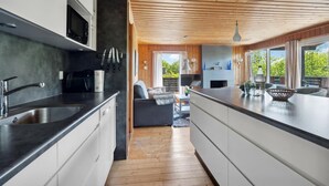 Ett modernt kök med en central köksö, träpanel i taket och stora fönster med utsikt över grönområden.