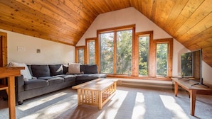 Ein gemütlicher Loftsraum mit schrägverdeckter Decke, einem Ecksofa, einem Holztisch und großen Fenstern mit Blick auf Bäume.