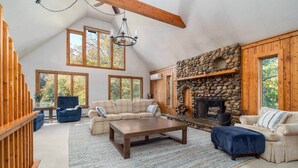 Ein geräumiges Wohnzimmer mit Kamin aus Stein, Holzbalken, großen Fenstern und einer gemütlichen Sitzecke.