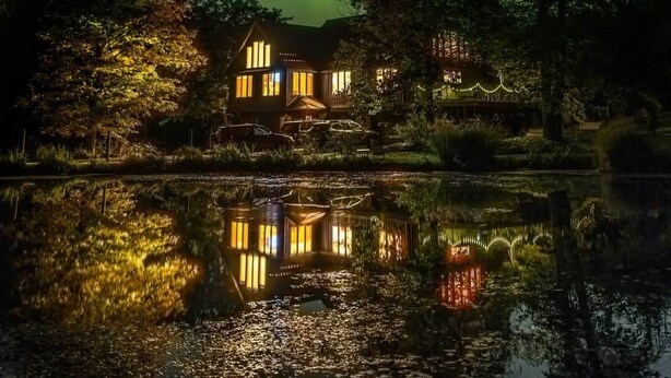 Ein Haus spiegelt sich in einem Gewässer, umgeben von Bäumen, unter einem sternenübersäten Nachthimmel.