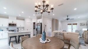 A modern kitchen with white cabinets, a central island, and a dining area with a round wooden table and woven chairs. - Sun-Kissed at Summer Salt (Orange Beach)