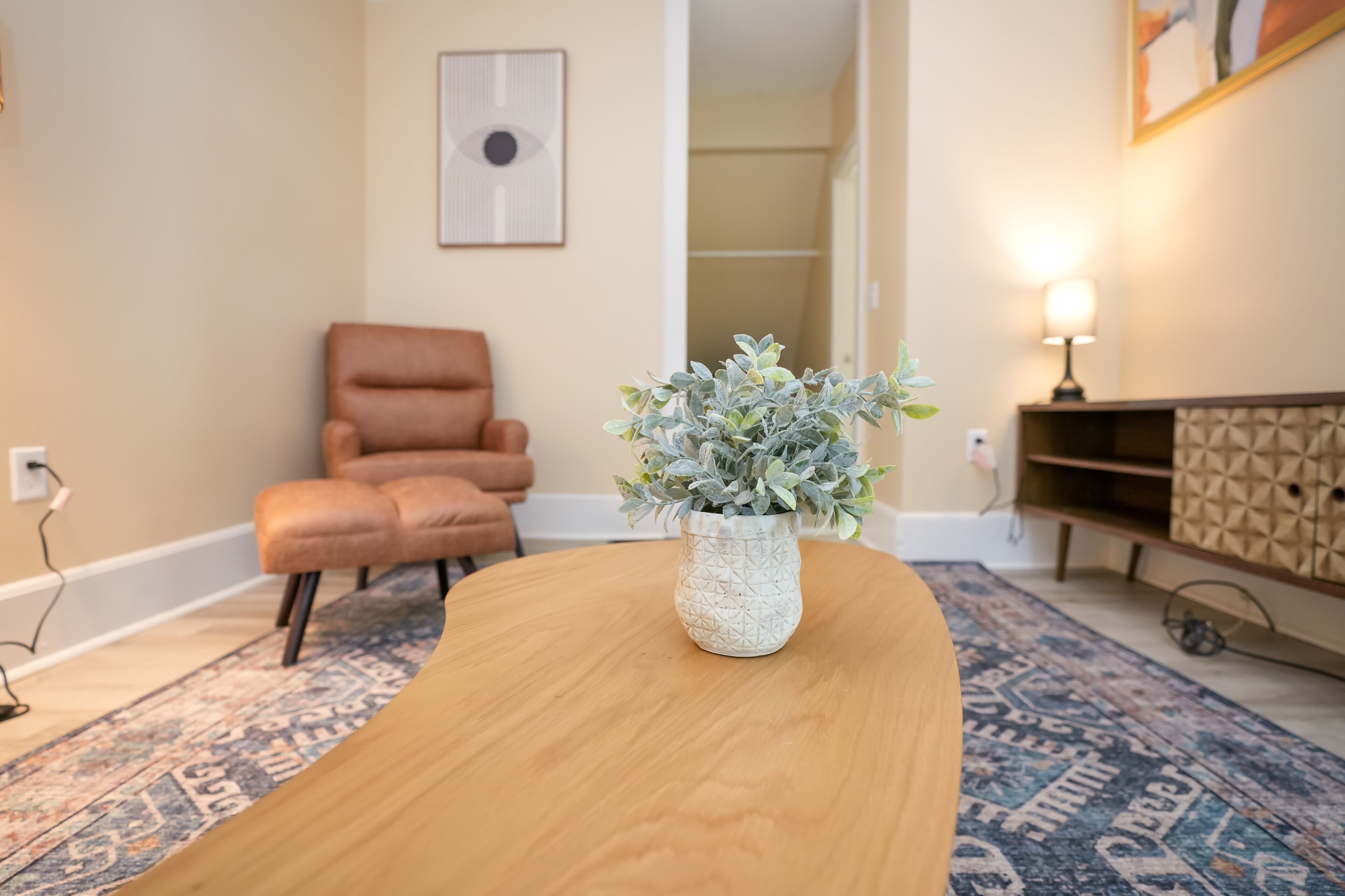 Quarto com uma mesa de madeira, um vaso com planta, uma poltrona de couro marrom, tapete estampado e uma obra de arte na parede.