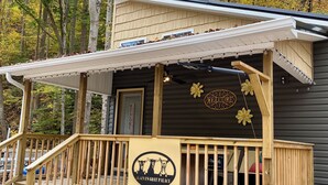 Une véranda en bois avec une enseigne indiquant « LA FERME DES CHÈVRES D’ELINA » et une pancarte de bienvenue au-dessus de la porte.