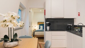 Una cocina moderna con armarios blancos, encimera negra y una máquina de café. Hay una zona de comedor con una mesa y sillas de madera, y una planta en maceta.