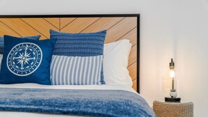 Una cama bien hecha con una almohada azul y blanca a rayas, una almohada azul con un diseño de brújula y una almohada blanca. Junto a la cama, una mesita de noche tejida con una lámpara.