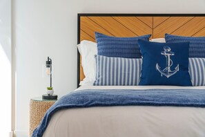 Een net opgemaakt bed met een blauw-wit gestreept kussen, een blauw kussen met een anker dessin, en een houten hoofdbord.