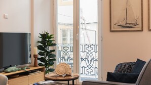 Una sala de estar con televisión, una planta, una mesa redonda de madera con un centro de troncos, un sofá y un balcón con barandilla.