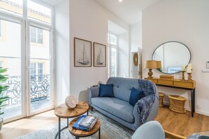 Een moderne woonkamer met een grijze bank, twee foto’s van zeilboten in lijsten, een ronde spiegel en een klein houten bijzettafeltje met boeken.