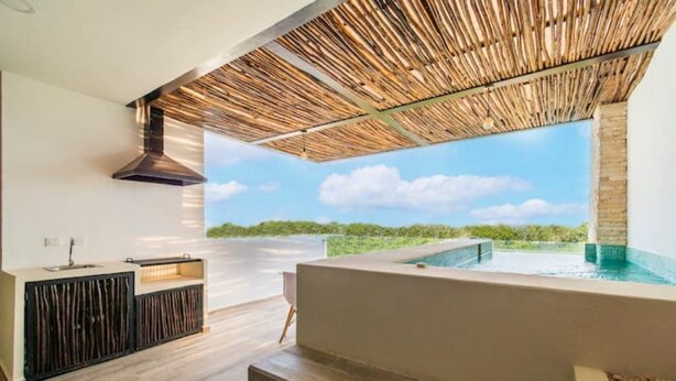 A modern outdoor kitchen area with a built-in barbecue, a large window overlooking a pool, and a stone wall. - Fabulous Apartment With Private Pool · 306 (Tulum)