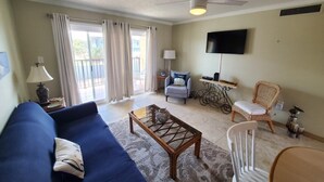 Un salon comprenant un canapé, une table basse et des fauteuils, un téléviseur fixé au mur, et un balcon avec des rideaux.