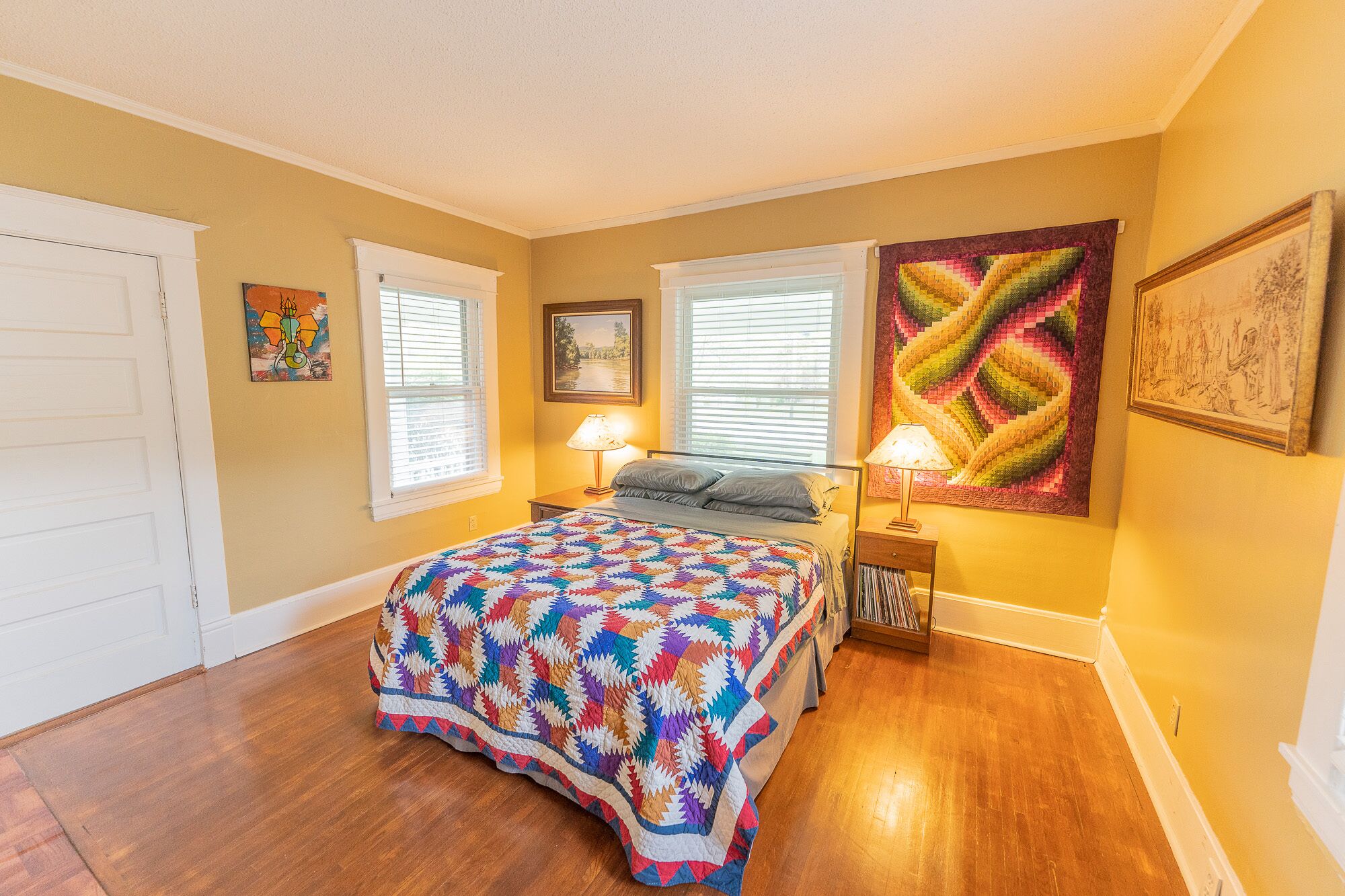 A bedroom with a wooden floor, a bed with a colorful quilt, two bedside lamps, a window with blinds, and a large quilted artwork on the wall.
