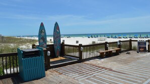 Eine Holzterrasse mit zwei Surfbrettern, die an einem Zaun lehnen, mit Blick auf einen Strand mit Sonnenschirmen und Menschen.