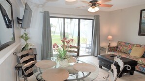 Ein Wohnzimmer mit einem Glastisch, umgeben von Stühlen, einer Couch und einem Wandfernseher, sowie einem Deckenventilator.