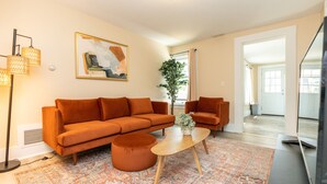 A living room with a sofa, ottoman, coffee table, and floor lamp. There is a painting on the wall and a plant in the corner. - Apartment in town center #1 (West Brookfield)