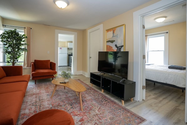 Una sala de estar moderna con un sofá, televisor y una mesita. Se ve una puerta que da a un dormitorio con cama.
