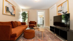 A living room with a sofa, ottoman, coffee table, TV stand, and artwork on the walls. - Apartment in town center #1 (West Brookfield)