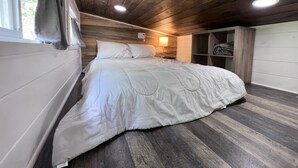 A bedroom with a wooden ceiling, a bed with white bedding, a nightstand, and a window with a curtain. - Unique Tiny Home Escape! Fully Equipped & Relaxing, Privacy-Fenced Outdoor Space (Raleigh)
