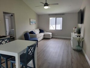A modern living room with a white sofa, a dining table with chairs, a ceiling fan, and a large window.