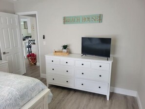 A bedroom with a bed, a dresser with a TV, a wicker basket, and a sign that says "Beach House".