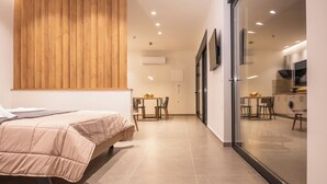 A modern bedroom with a bed, a wooden panel wall, a small dining area, and a kitchen visible through a glass door.