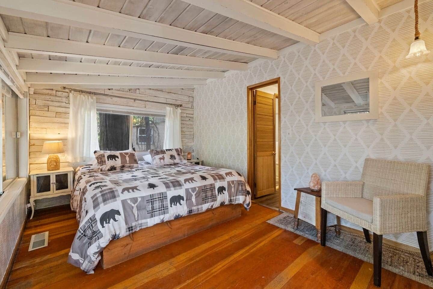 A bedroom with a wooden floor, a bed with a patterned bedspread, a chair, and a window with curtains.
