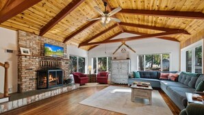 A spacious living room with a fireplace, wooden floors, a stone accent wall, and large windows. - Walk to town, Lake access, Pet Friendly, King bed (Twain Harte)