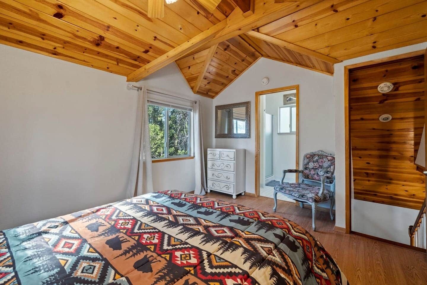 Une chambre avec un plafond en bois incliné, un lit recouvert d’une couverture à motifs, une commode, une chaise et un miroir accroché au mur.